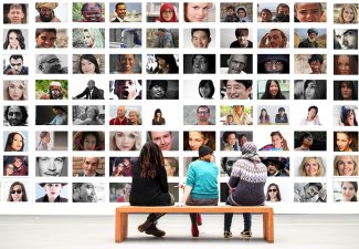 Image of three people sitting on a bench in front of a wall featuring the faces of many different people