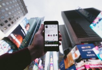 Photo of a person holding up a phone with the Instagram app opened with the backdrop of many buildings