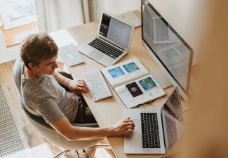 Photo of a person with two laptop computers on either side of them, an open book in front of them, and a desktop computer in front of them