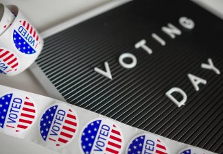 A close-up image of a blackboard with the words “VOTING DAY” spelled out in white capital letters. In the foreground, there is a roll of circular stickers with an American flag design and the text “I VOTED” prominently displayed. 