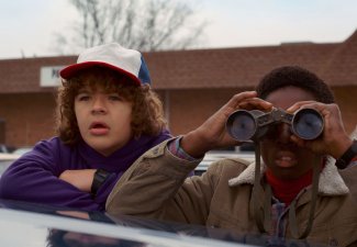 Still from Stranger Things show with two characters observing something in distance, one has binoculars