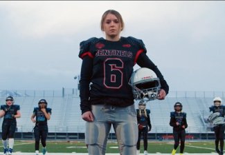 Young girl in football uniform standing in a field, looking at the camera as other girls train behind her.