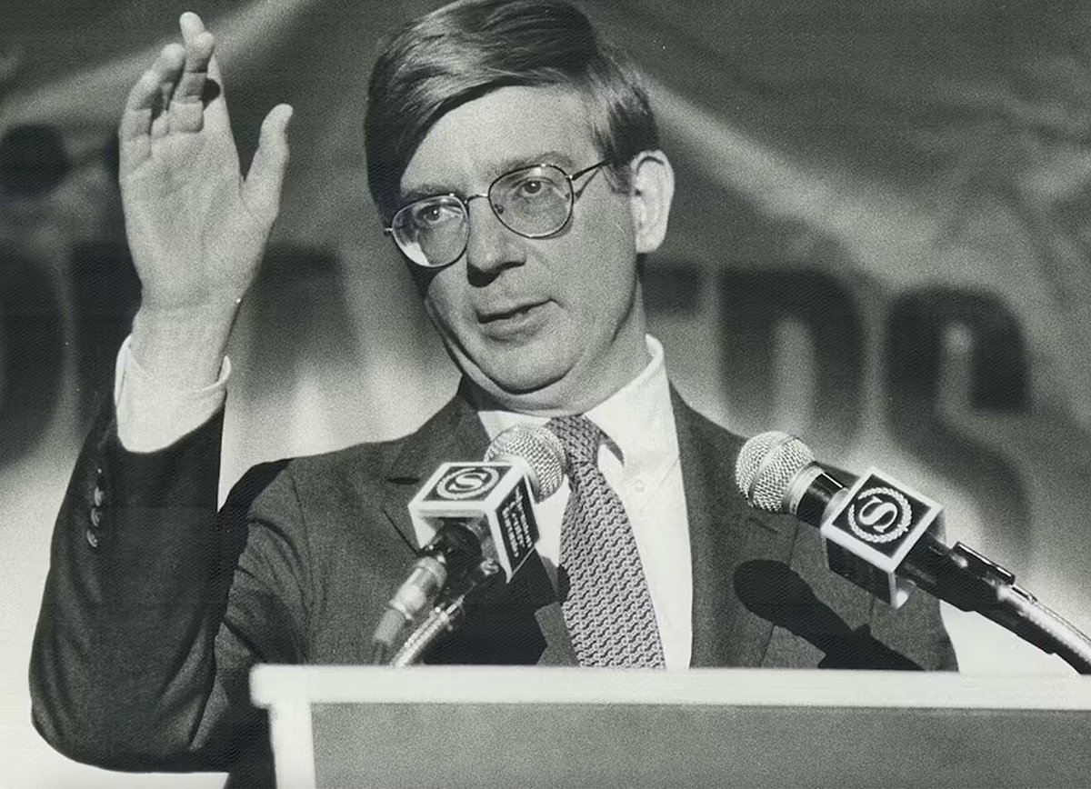 Black and white photo of Washington Post columnist George Will stands in front of a podium with his hand raised.