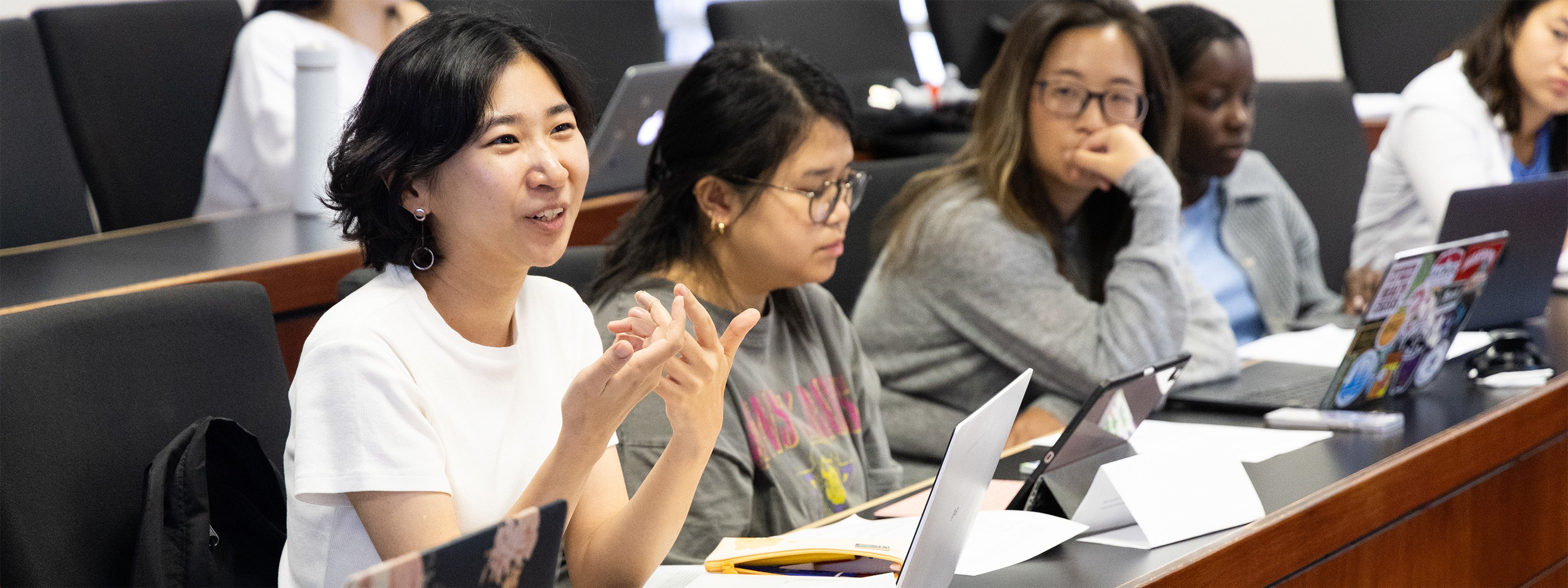 A classroom setting with students seated in rows, each with laptops or notebooks on desks. The focal point is a woman in a white shirt actively participating in a discussion, gesturing with her hands. Other students appear attentive.
