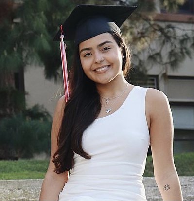 Jenisty Colon stands in front of building with a white dress and black graduation cap on, celebrating her graduation from Chapman University.