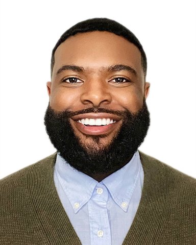 bearded Black man with green sweater over blue shirt smiles for camera in front of white background