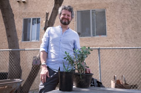man in blue button up shirt and grey pants smiles for camera while holding two plants