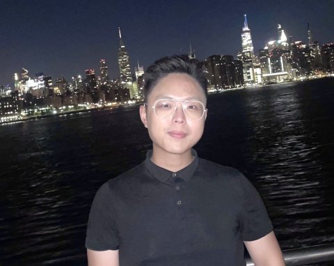 Male Asian man wearing clear framed glasses stands in front of Taipei skyline