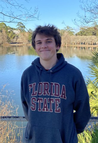 Jack Brockman, a new freshman at USC stands in front of a blue lake with green forage; bushes and brown branches.