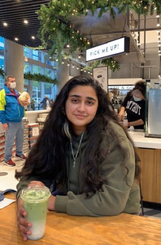 A woman named Raka Bhattacharyya sits at a shop holding a smoothie in her hand and smiling at the camera.