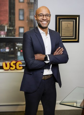 Anthony Galloway stands in front of USC sign and plaque while smiling with arms folded. 