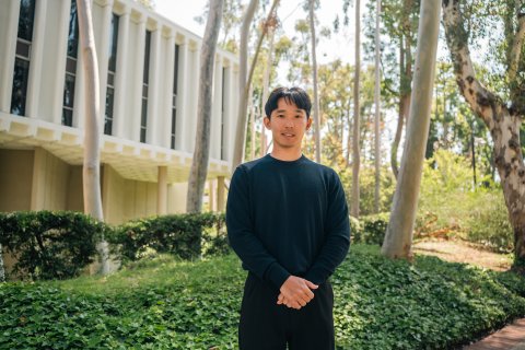 Anthony Moon stands in front of greenery and trees in front of the white Annenberg School for Communication building, smiling and wearing all black with his hands folded in front of him.