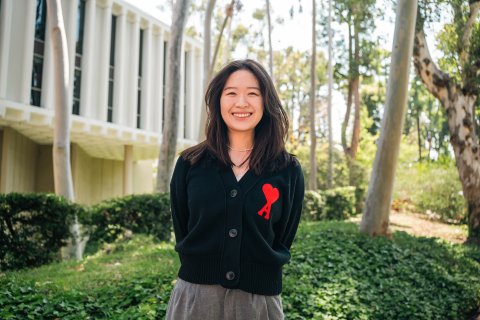 Ruby Chang stands in front of green foliage and the white USC Annenberg School for Communication Building, wearing a black cardigan with a red A and red heart on the front.