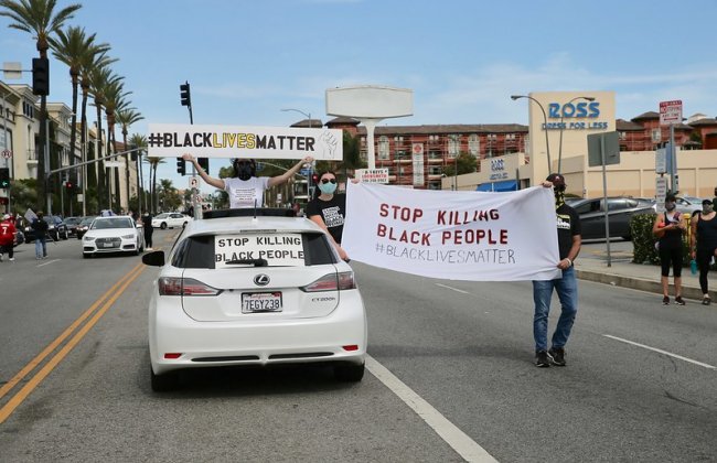 Photo of a Black Lives Matter protest on the street where a person sits on top of a car with a sign that says #BlackLivesMatter