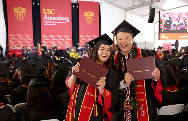 Two individuals at a graduation ceremony, wearing black caps and gowns with red stoles and holding up their diplomas, which have ‘University of Southern California’ written on them. They are standing in front of seated graduates and a stage