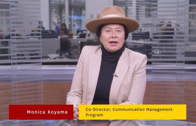 Still from a video in which Monica Koyama, wearing a tan coat and a wide brimmed hat, discusses the Comm Management program from inside a studio in the Media Center.