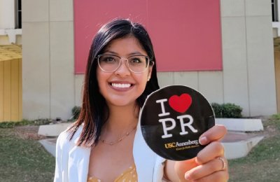 USC Annenberg public relations student Dany Rodríguez Martinez holds a black "I Love PR" button in front of a building.