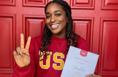 black female college student wearing red sweater holds up victory hand sign while showing acceptance letter in front of red door