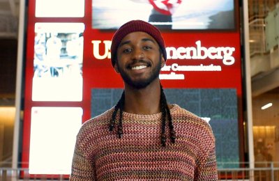 Black male in red beanie hat smiles in front of USC Annenberg media wall