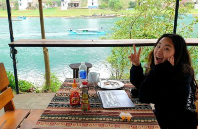 women smiles for camera at picnic table with computer with coastal Thailand in the background. 