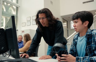 A student with glasses and a black shirt bends over a computer desk to help another student with his work.