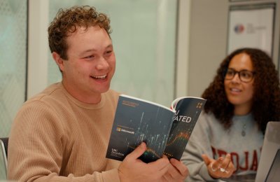 A man wearing a tan shirt holds a research report as another student looks on.