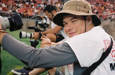 male in tan bucket hat smiles at sporting event holding camera