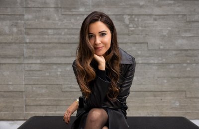 Woman wearing all black sits on a bench in front of a stone wall. She is leaning forward with her elbow on one knee, smiling slightly. 