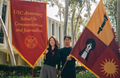 Two individuals stand in front of the Annenberg School for Communication Building holding flags. On the left, a woman holds a cardinal flag bearing the name of the school, and on the right, a man holds a flag bearing symbols that represent the school.