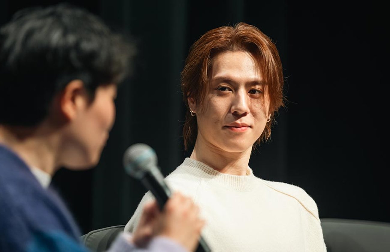 Yugyeom, wearing a white shirt and sitting on a stage with a black background, listens as Clinical Assistant Professor of Communication Hye Jin Lee, facing away from the camera wearing a blue shirt and holding a microphone, asks a question.
