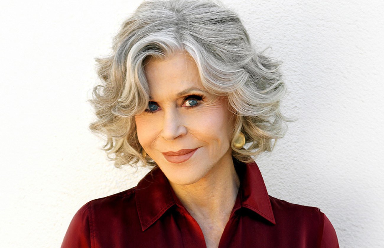 Activist and actor Jane Fonda smiles at camera, wearing a red collared shirt against a white background. 