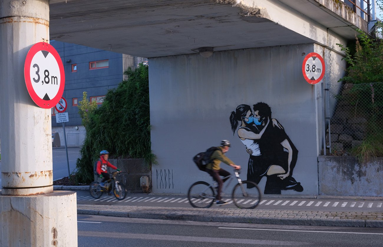 Two people on bicycles under a highway that has an illustration painted on of two people kissing with medical masks on