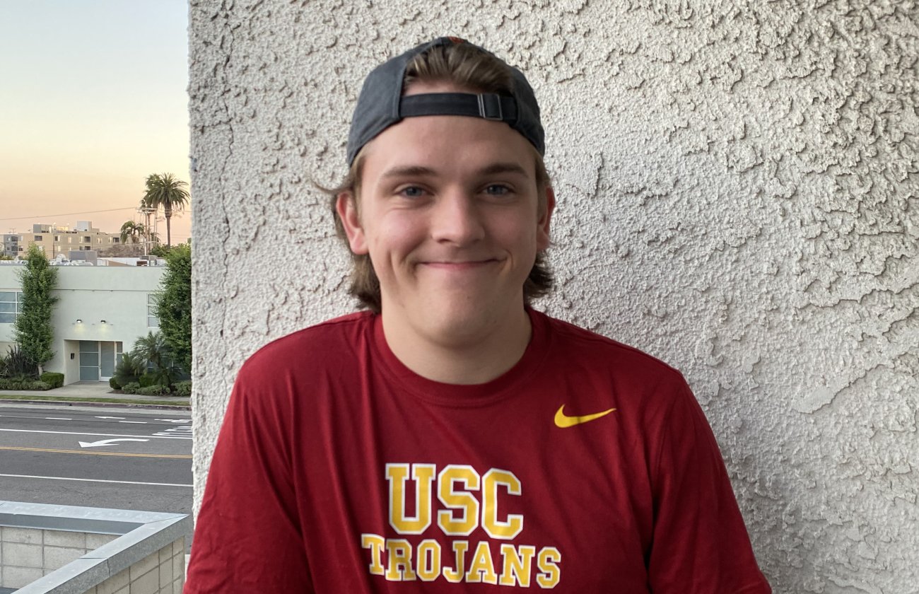 Jack Brockman, a new freshman at USC stands against the wall with a red and gold-lettered USC Trojans shirt and black backwards cap.