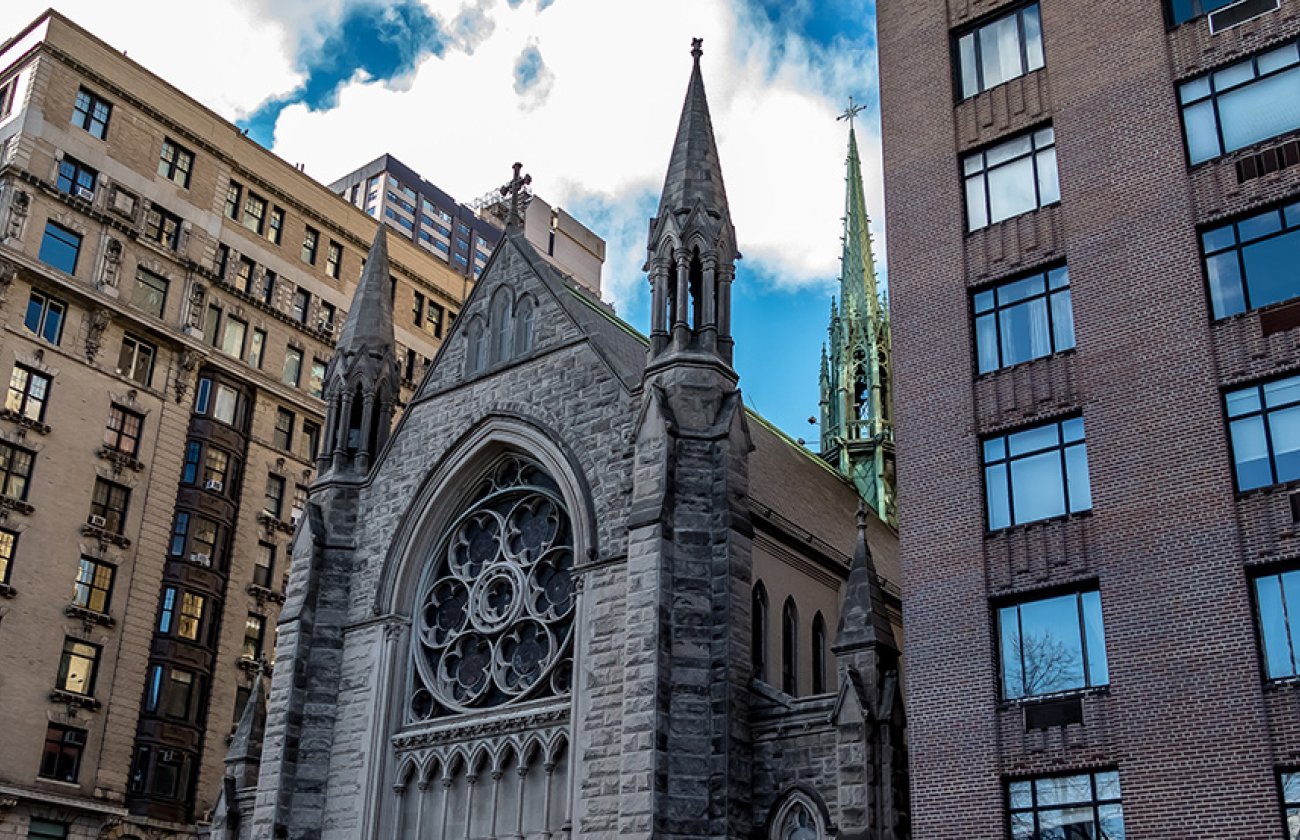 A 3/4ths view of a cathedral building sandwiched between two larger metropolitan buildings on a sunny day