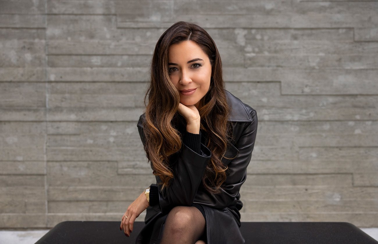 Woman wearing all black sits on a bench in front of a stone wall. She is leaning forward with her elbow on one knee, smiling slightly. 