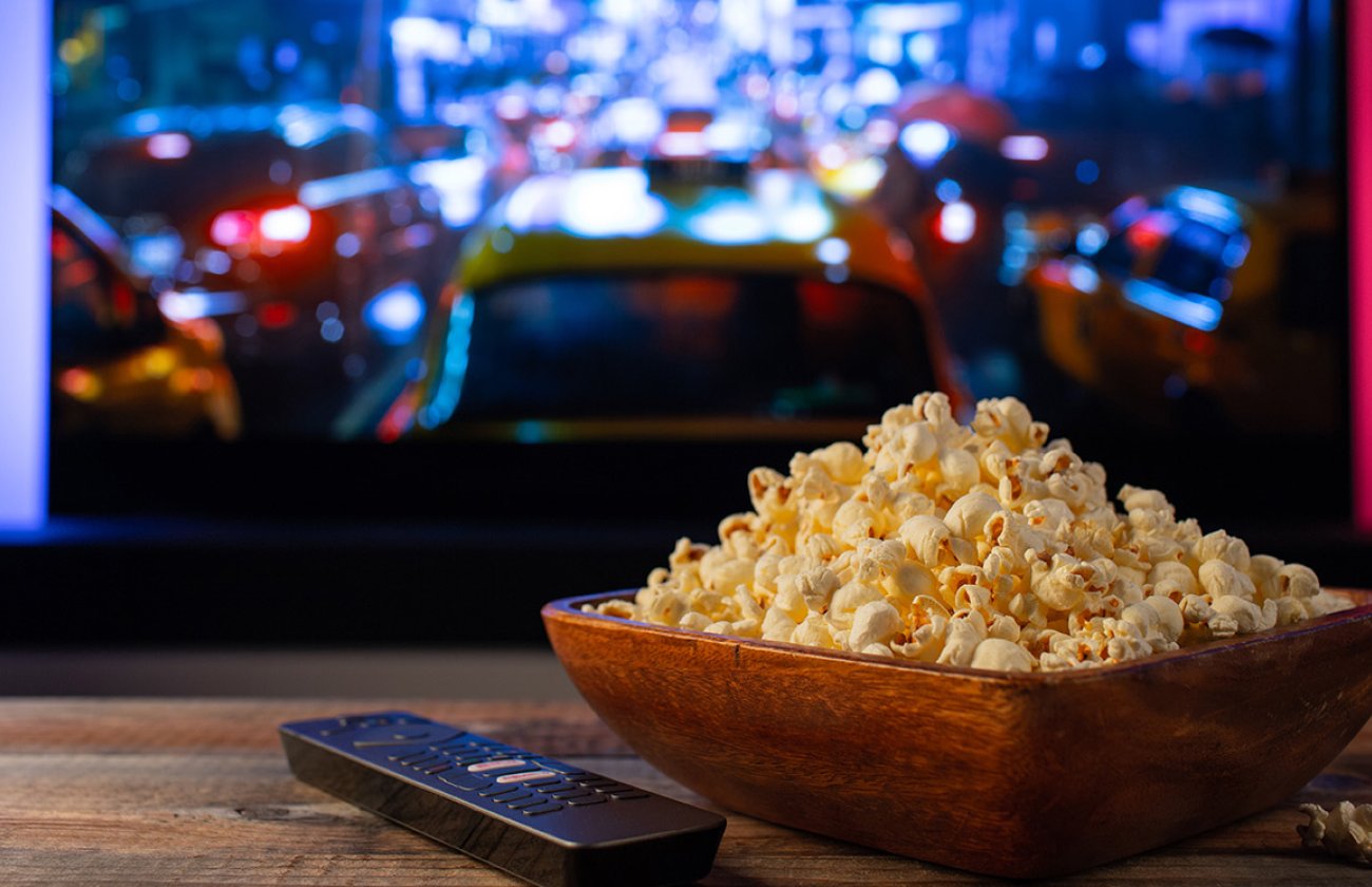 A bowl of popcorn sits on a coffee table with a remote next to it in front of a TV.