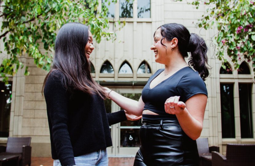 female Asian college student wearing black sweater and jeans laughs with female Latinx student in black shirt and pants