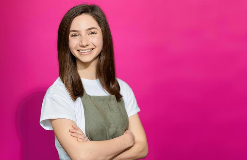woman in early 20s wearing cooking apron poses in front of pink background