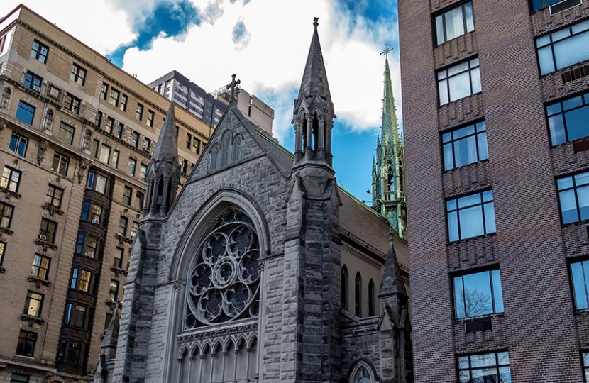 A 3/4ths view of a cathedral building sandwiched between two larger metropolitan buildings on a sunny day