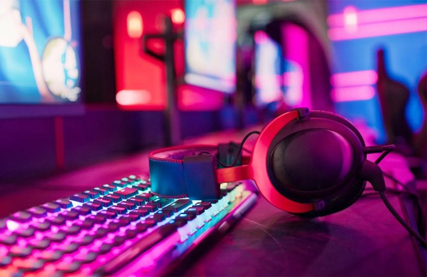 A set of headphones rests on a keyboard, which is in front of a computer screen that displays a video game. The lights are colorful and evoke cyber space. 