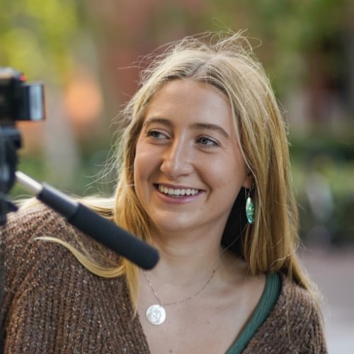 Young woman standing in front of a video camera propped up on a tripod. She appears to be filming something in front of a USC building.