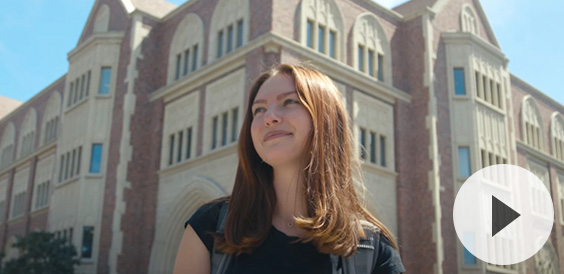 A young woman with shoulder-length brown hair standing in front of a grand brick-and-stone academic building under a bright blue sky, smiling and looking upwards.