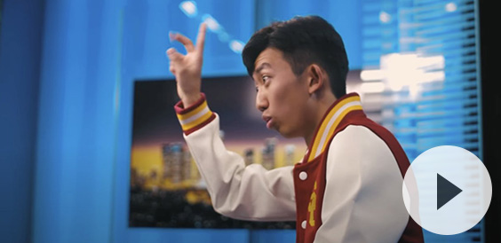 A young man in a cardinal, gold and white varsity jacket animatedly gesturing with his hand while speaking. He is indoors, with a blurred cityscape image in the background.