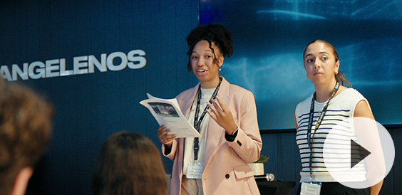 Two women presenting in front of an audience. One holds a booklet while speaking, and the other listens attentively. Behind them, the word 