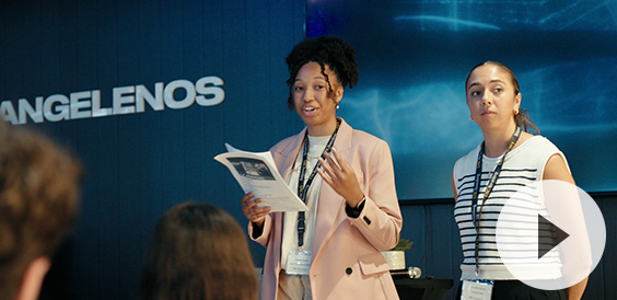 Two women presenting in front of an audience. One holds a booklet while speaking, and the other listens attentively. Behind them, the word 'ANGELENOS' is visible on the wall.