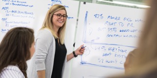 Female smiling and pointing at a whiteboard behind her.